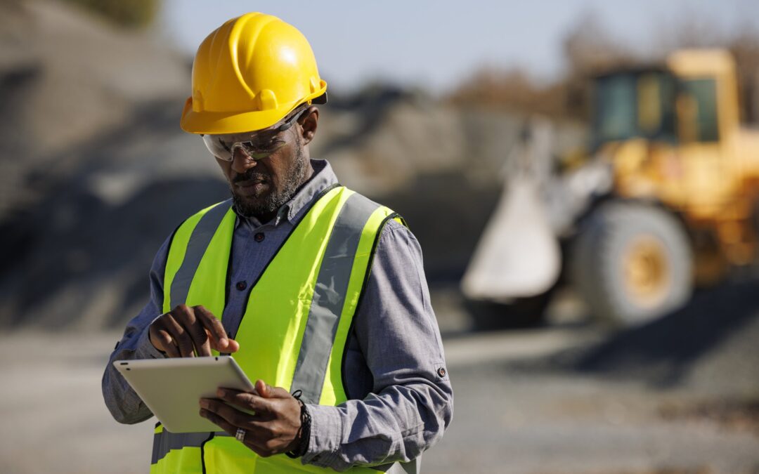 African American miner with mining career working at job site in 2024 wearing hard hat and looking at tablet with bulldozer in background