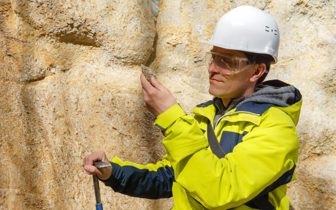 Male geologist in helmet and protective glasses examines a sample of the mineral outdoors against a rock for mining industry
